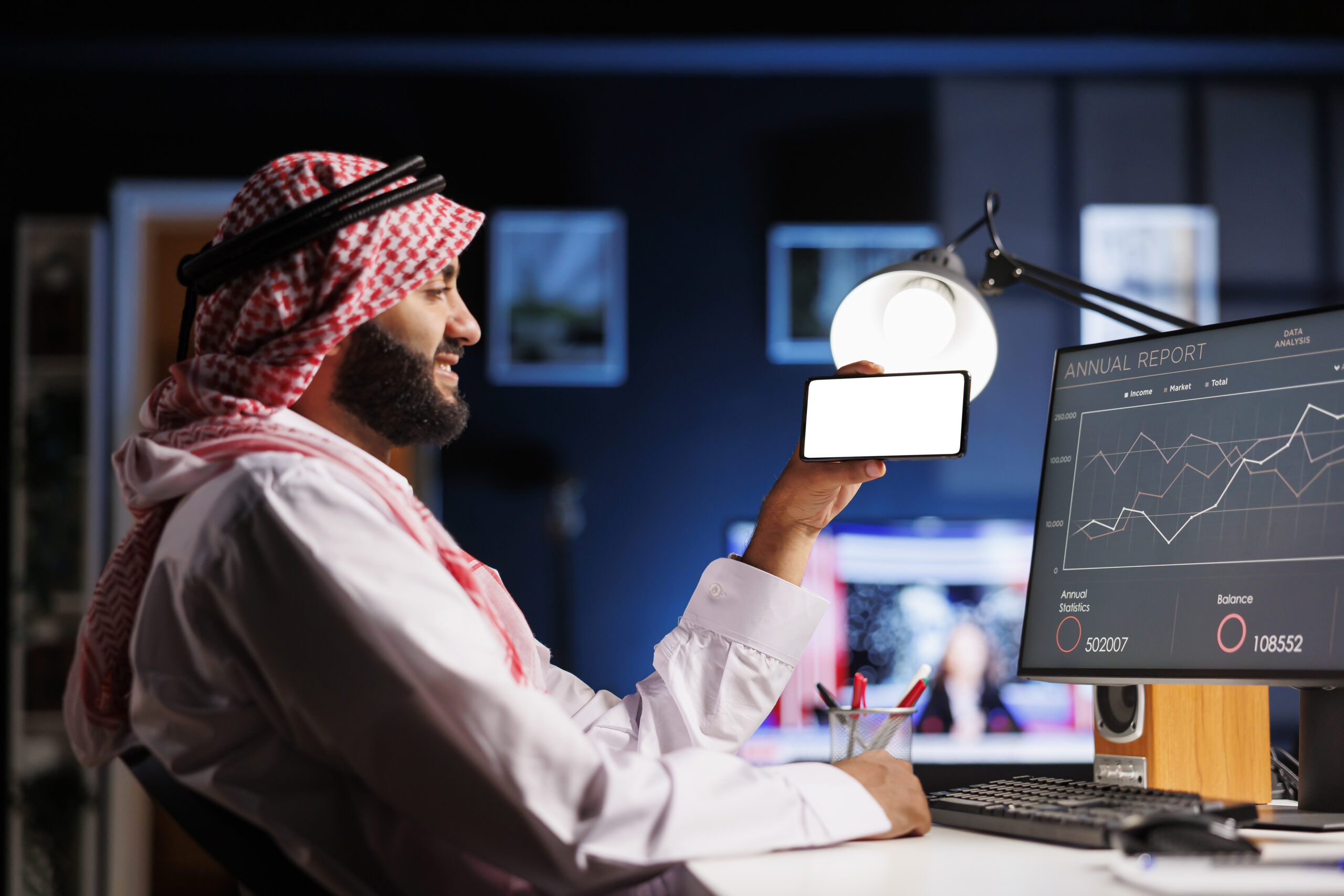An Arabic individual, wearing traditional clothing uses a mobile phone at an office desk. The isolated white screen offers space for customized content in an advertising or promotional setting.